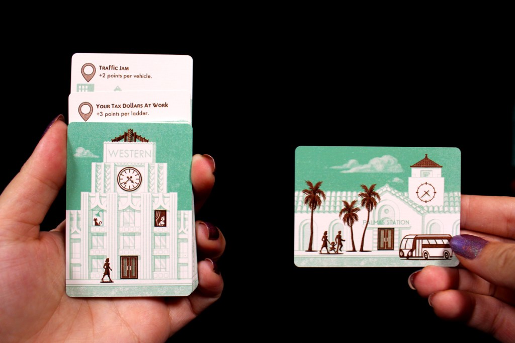 A hand holding two game cards. The card on the left features a building labeled 'WESTERN' with a clock and illustrations indicating points for 'Traffic Jam' and 'Your Tax Dollars At Work.' The card on the right shows 'PALMS STATION' with palm trees, a bus, and pedestrians.