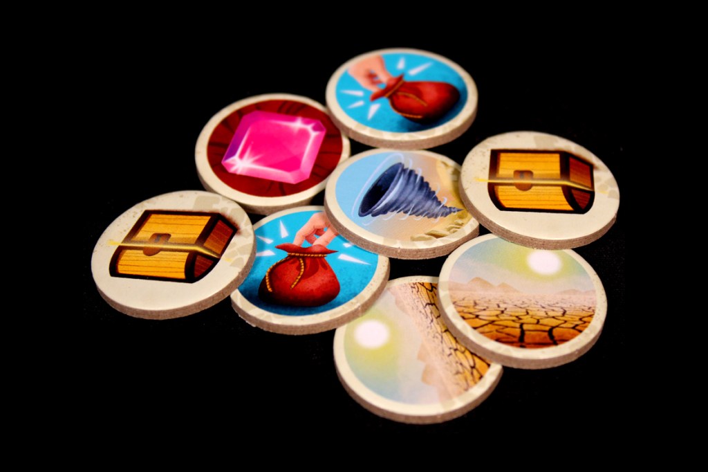 A collection of round game tokens featuring various illustrations such as a gem, a treasure chest, a hand holding a bag, and an image of a drought landscape, all set against a black background.