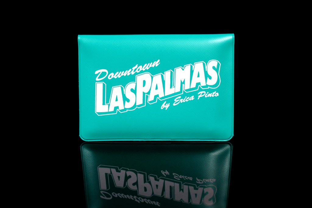 A teal clutch featuring the words 'Downtown Las Palmas by Erica Pinto' in bold white lettering.