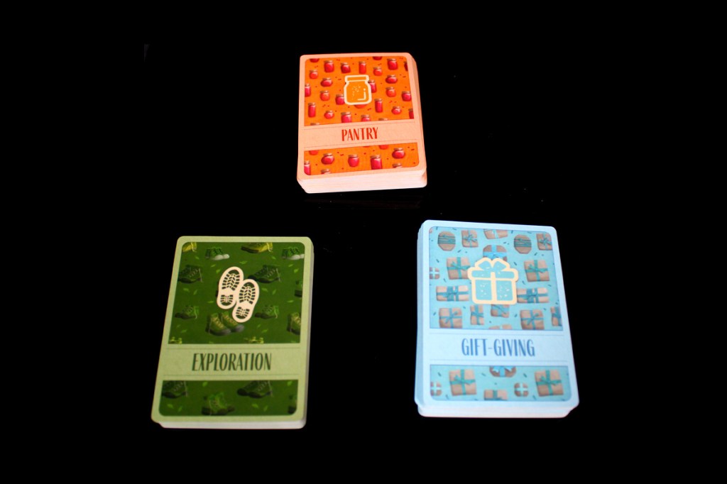 A collection of three colorful game cards on a black background, titled 'Pantry' (orange), 'Exploration' (green), and 'Gift-Giving' (blue).