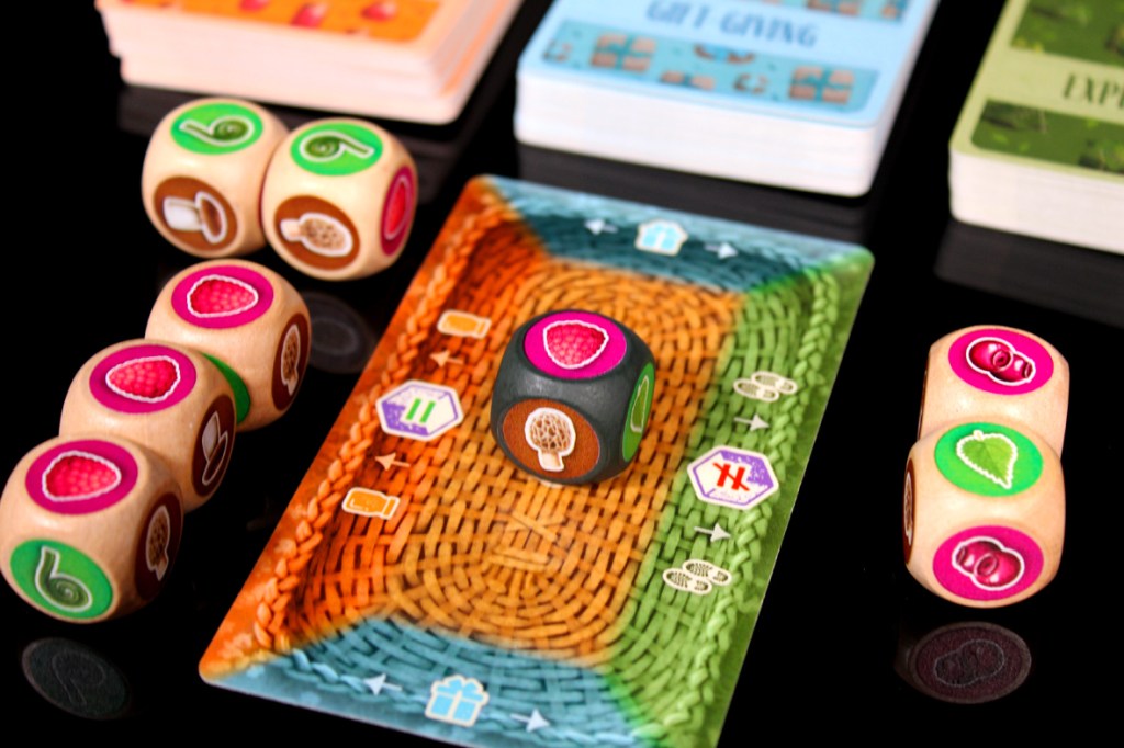A colorful game board with fruit-themed dice, including pink and green designs, along with several card decks in the background.