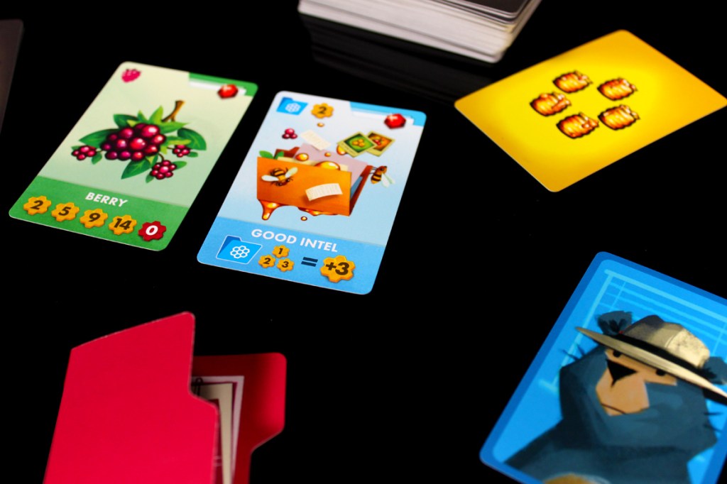 A close-up view of several colorful game cards on a black surface, including a card labeled 'Berry' with berry illustrations and statistics, a card labeled 'Good Intel' featuring a playful design with math symbols, and another card displaying a bear character wearing a hat.