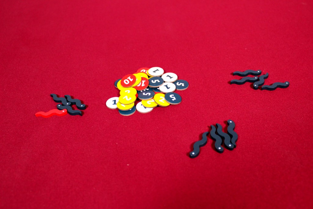 A pile of colorful game tokens and several dark wavy plastic pieces scattered on a red tablecloth.