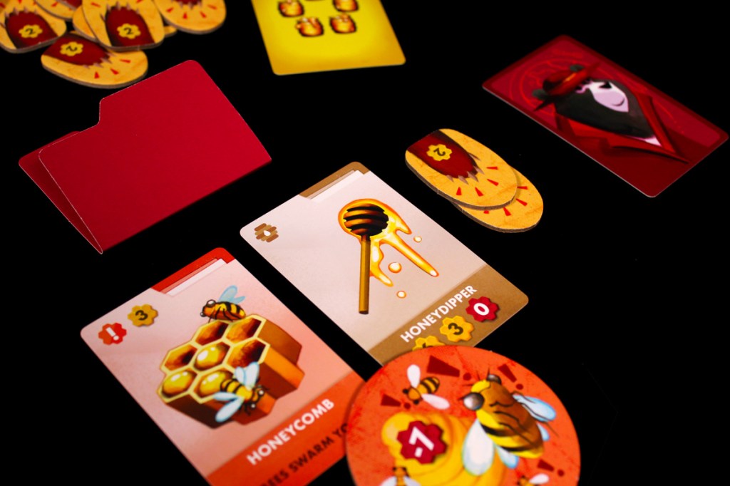 A flat lay of various game cards on a black background, featuring illustrations of honeycomb, a honey dipper, and buzzing bees, along with a red folder.