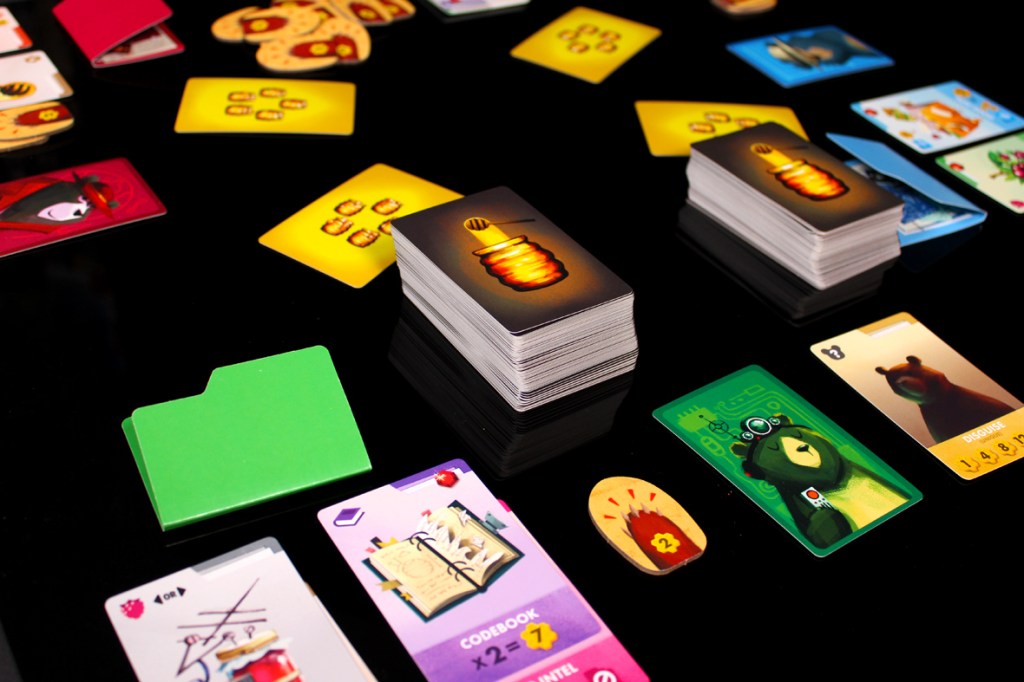 A tabletop game setup featuring various playing cards, including character cards, action cards, and a green folder. The game elements are displayed on a black surface, highlighting numerous colorful cards and tokens related to gameplay.