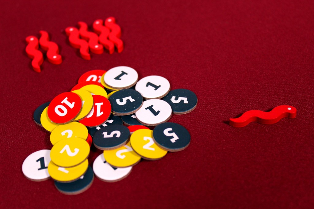 A collection of colorful game tokens and pieces on a red surface, including round tokens with numbers and a red worm-shaped piece.