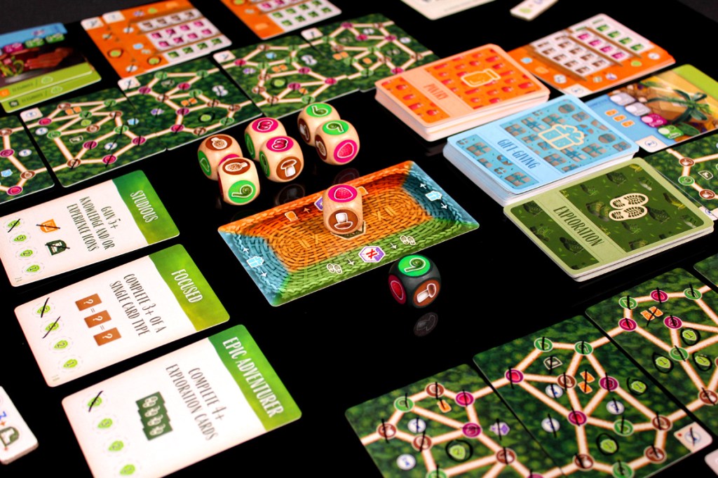 A tabletop game setup featuring various cards, dice, and game boards with distinctive colors and designs, displaying actions, goals, and game mechanics.