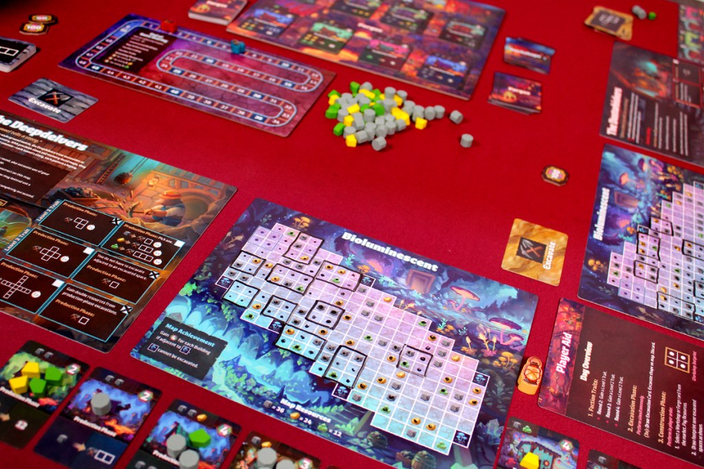 A tabletop game setup featuring various game boards, player aids, and colorful gaming pieces on a red tablecloth.