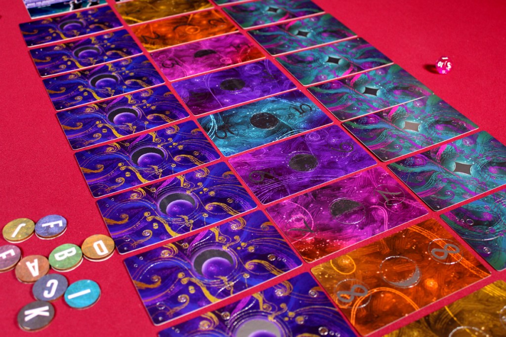 Colorful game cards arranged in rows on a red table, accompanied by tokens with letters and a pink die.