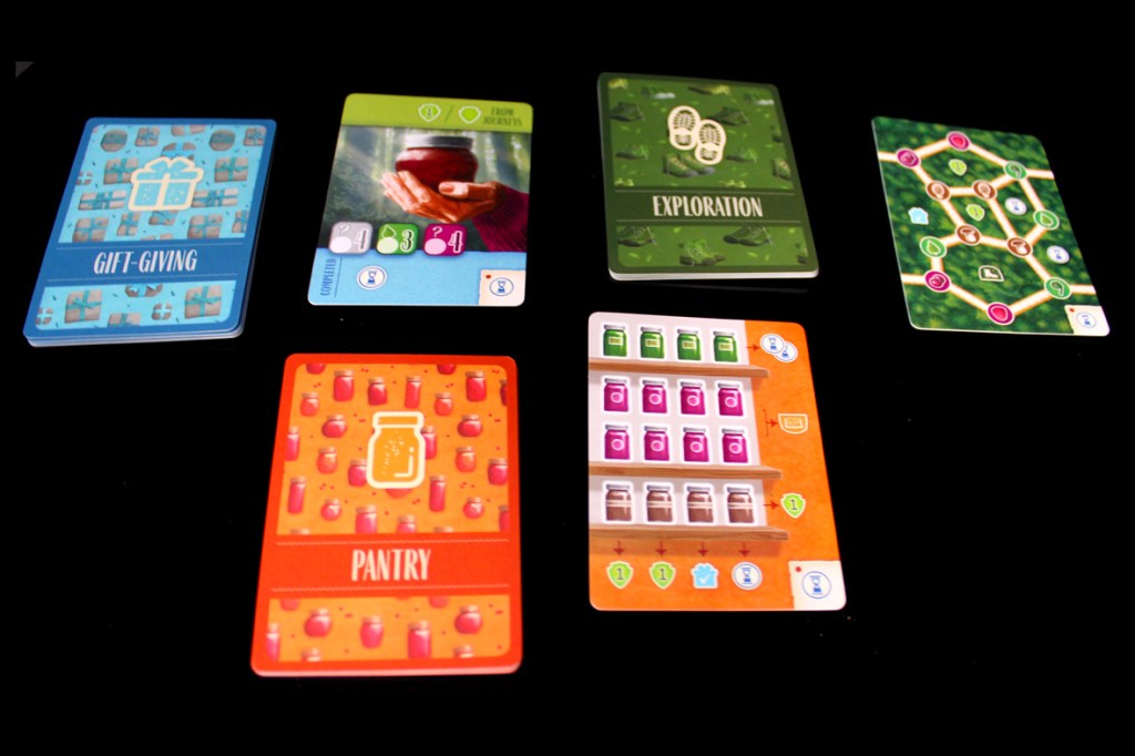 A flat lay of six colorful game cards on a black background, including cards labeled 'Gift Giving,' 'Exploration,' and 'Pantry,' each featuring distinctive designs and icons.
